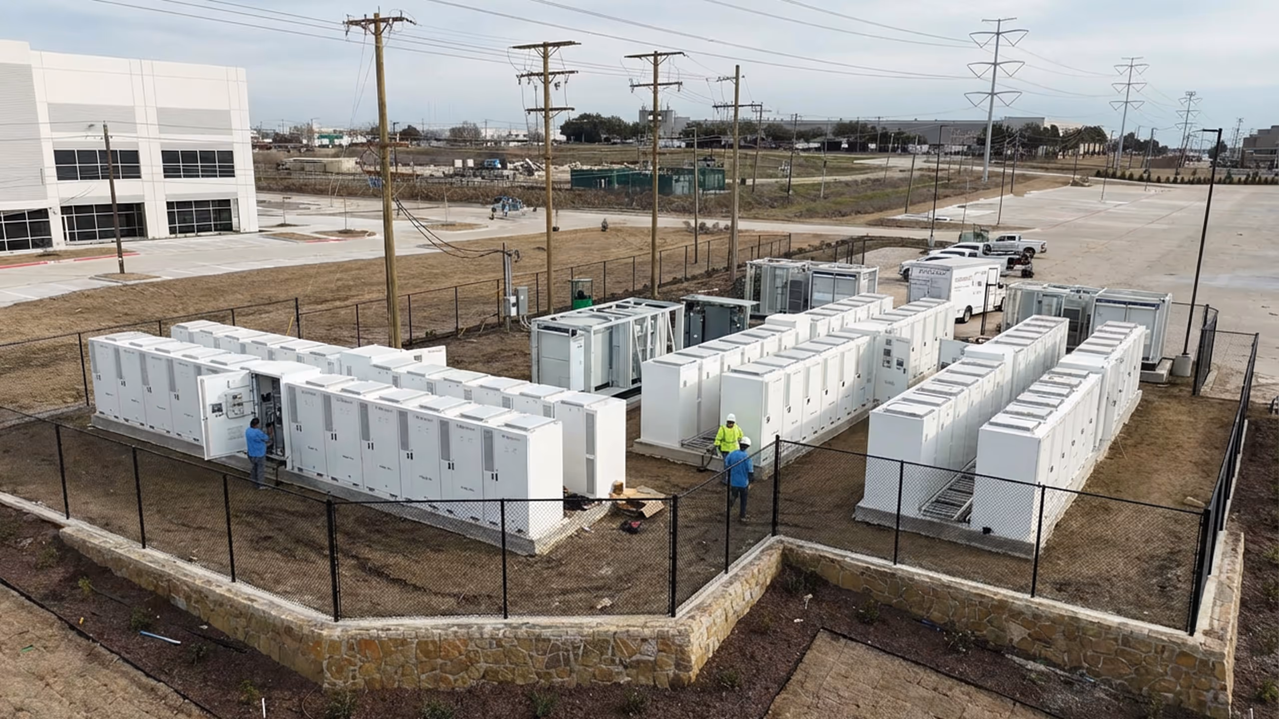 Fenced-in outdoor area with multiple rows of large white electrical cabinets and two workers in safety gear.