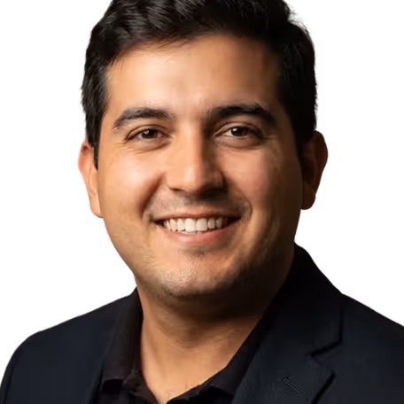 Smiling man with dark hair wearing a black blazer and shirt against a black background.