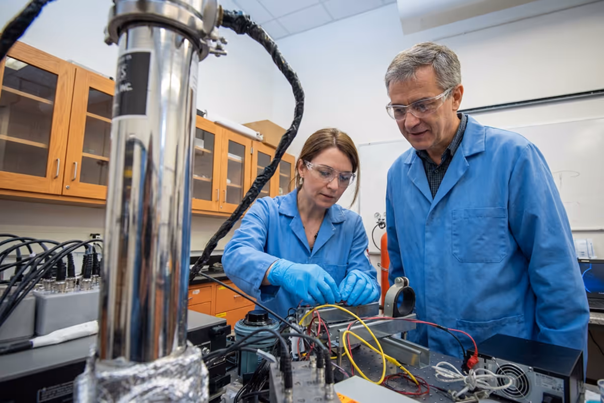 Two scientists in blue lab coats and safety glasses working with electronic equipment and wires in a laboratory.