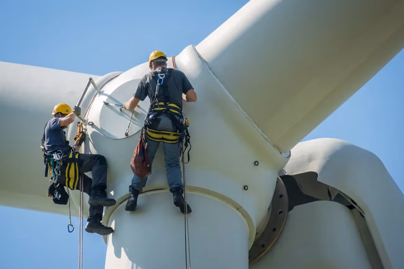 Two workers wearing helmets and safety harnesses performing maintenance on a wind turbine against a clear blue sky.