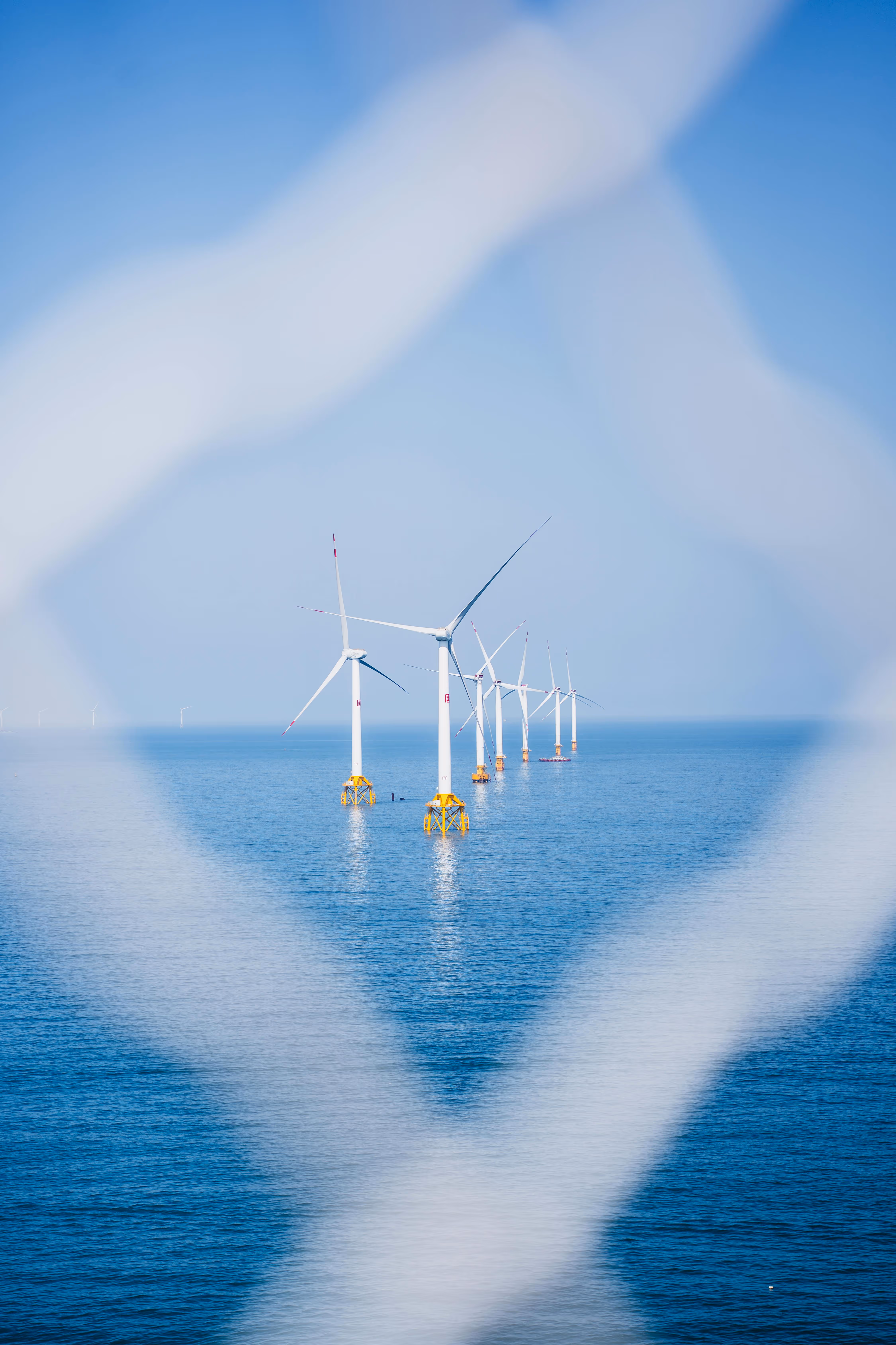 Offshore wind turbines with yellow bases arranged in a line in blue ocean water, viewed through a diamond-shaped metal fence.