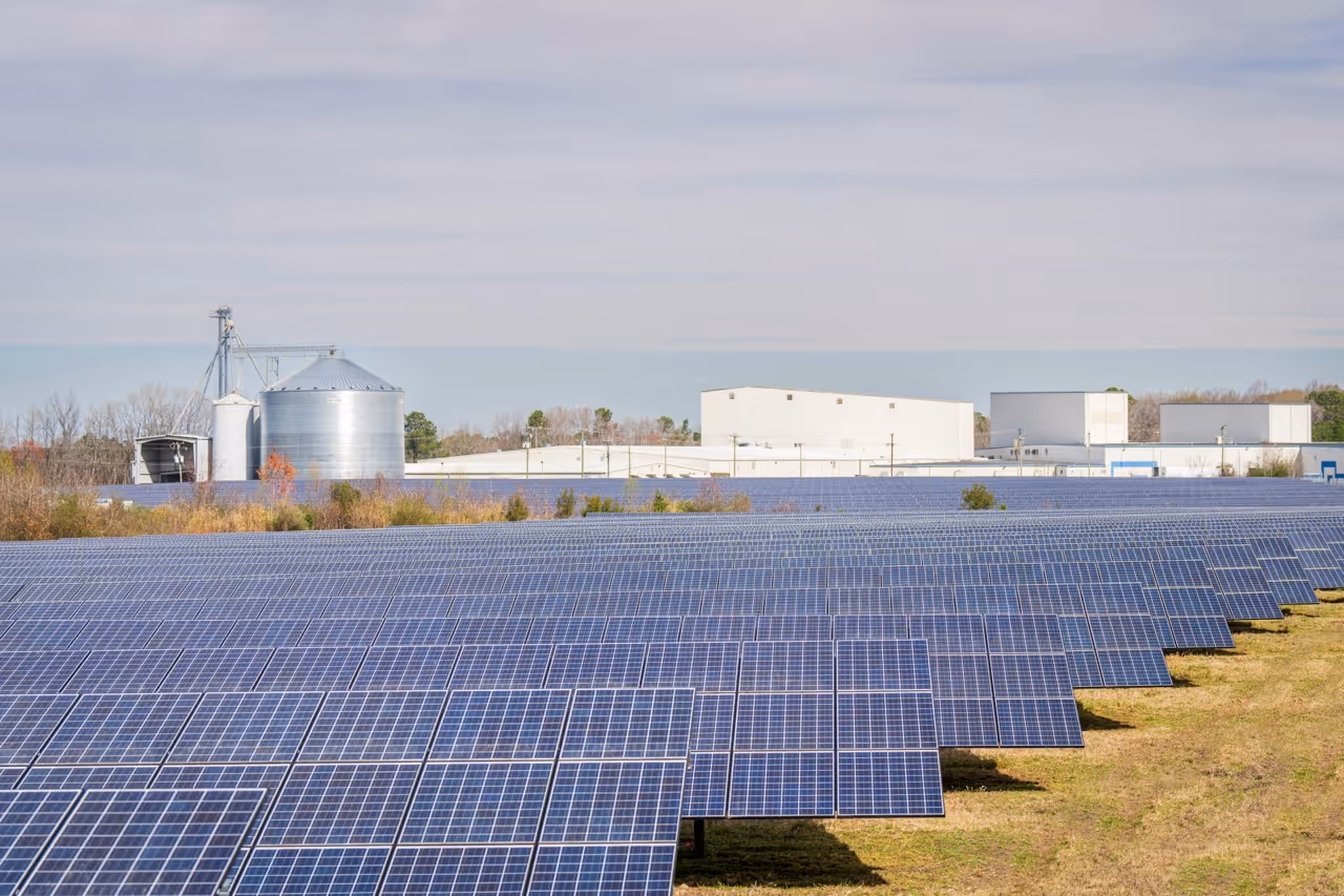 Large solar panel farm with rows of solar panels in front of industrial buildings and silos on a partly cloudy day.