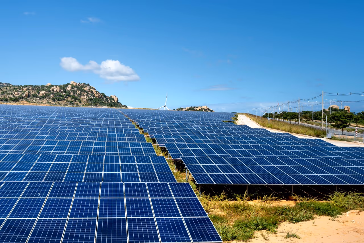 Large solar panel farm under clear blue sky with hills and wind turbines in the background.