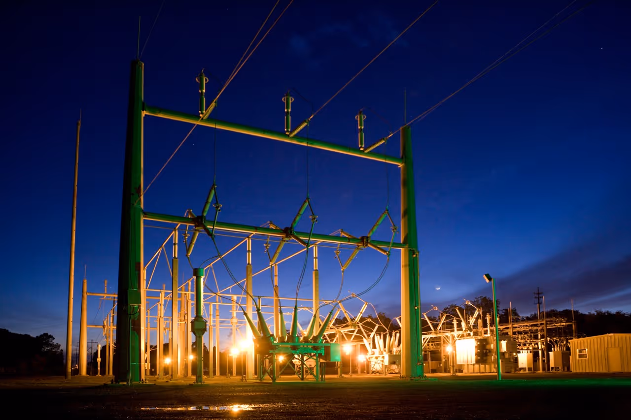 Electrical substation with illuminated transformers and power lines under a deep blue evening sky.